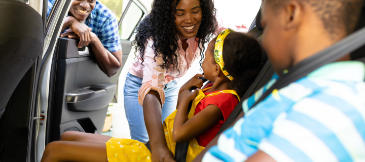 Smiling African American mother teaching daughter to wear seat belt while sitting in car.